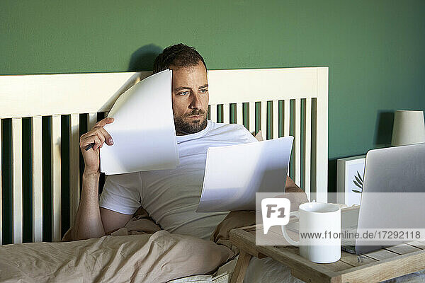 Mid adult man with document looking at laptop while resting in bed