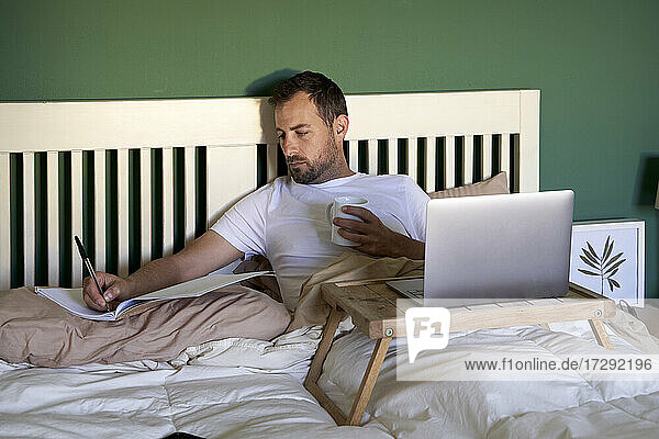 Mann mit Kaffeetasse  der in ein Buch schreibt  während er zu Hause im Bett arbeitet