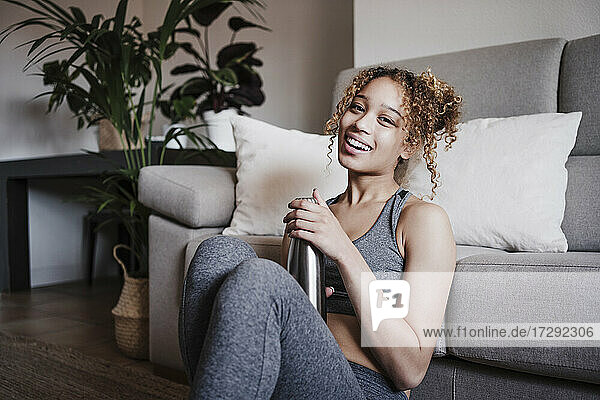 Smiling curly haired woman with bottle sitting in front of sofa at home