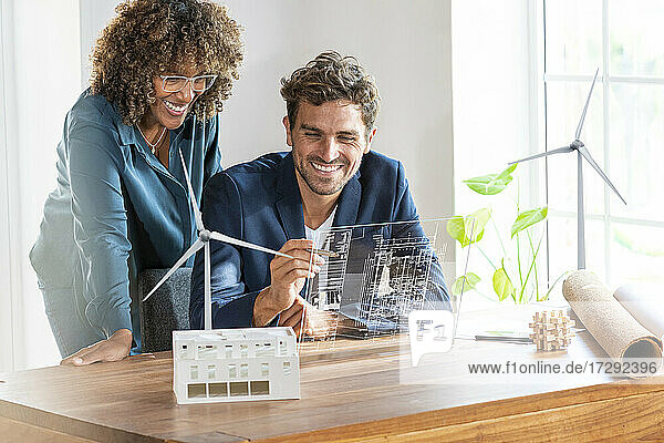 Kollegen im Büro schauen auf einen futuristischen Bildschirm mit einem Architekturplan eines nachhaltigen Hauses und Windturbinen