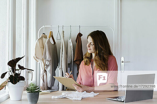 Weibliche Modedesignerin schreibt in ein Klemmbrett  während sie zu Hause im Büro sitzt