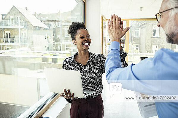 Fröhliche Geschäftsfrau mit Laptop  die mit einem männlichen Kollegen im Büro high five macht