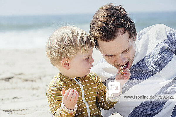 Son feeding snack to father while sitting at beach
