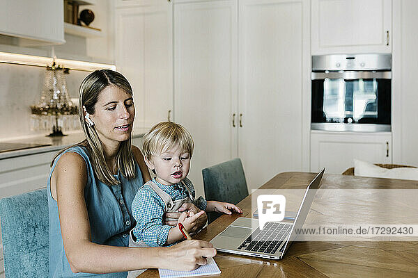 Mittlere erwachsene Frau  die während eines Videoanrufs in ihr Tagebuch schreibt  während ihr Sohn zu Hause auf dem Schoß sitzt