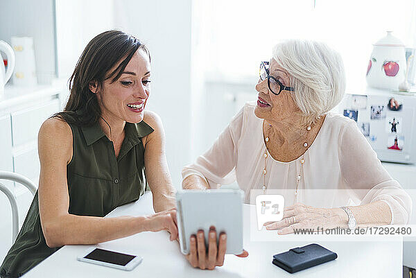 Ältere Frau im Gespräch mit ihrer Enkelin  die ein digitales Tablet benutzt  während sie am Tisch sitzt