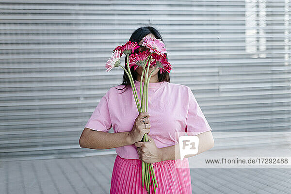 Junge Frau bedeckt ihr Gesicht mit Gerbera-Gänseblümchen vor einem Wellblechdach