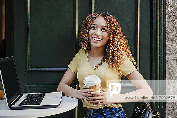 Smiling young woman with laptop holding reusable cup while sitting at sidewalk cafe