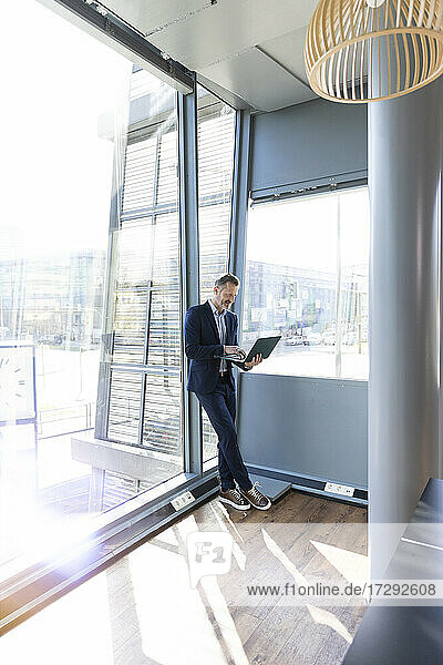 Männlicher Berufstätiger arbeitet mit Laptop und lehnt sich an ein Glasfenster im Büro