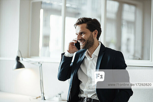 Lächelnder Geschäftsmann  der im Büro mit seinem Smartphone spricht