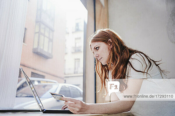 Rothaarige Frau bei der Online-Zahlung per Kreditkarte  während sie einen Laptop vor einem Fenster zu Hause benutzt