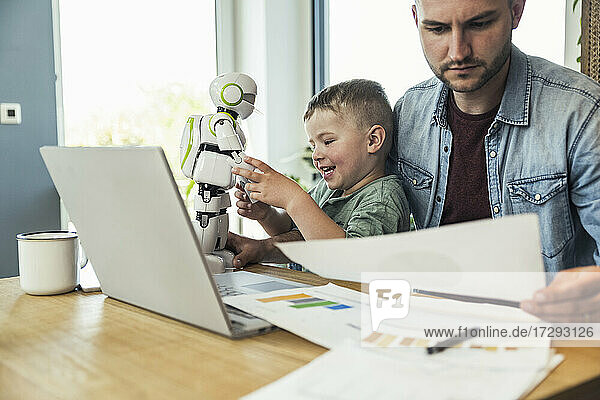 Lächelnder Junge spielt mit einem Roboter  während ein Geschäftsmann zu Hause Dokumente liest