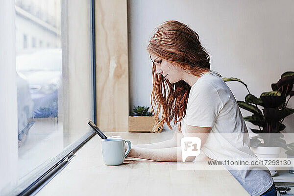 Redhead woman using mobile phone while leaning on window sill at home