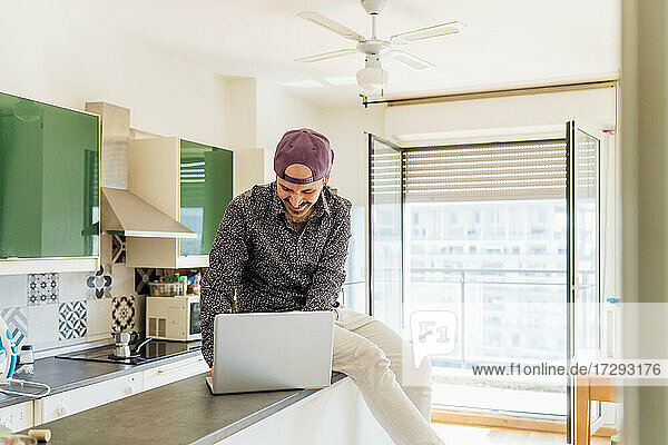 Lächelnder junger Mann  der einen Laptop benutzt  während er auf einer Kücheninsel zu Hause sitzt
