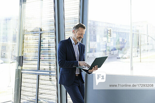 Lächelnder Geschäftsmann arbeitet am Laptop vor dem Fenster im Büro