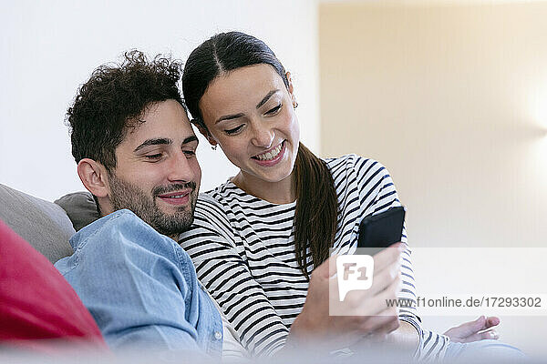 Lächelndes Paar  das im heimischen Wohnzimmer ein Mobiltelefon benutzt