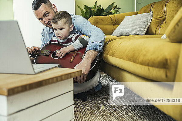 Junge  der Gitarre spielen lernt  sitzt mit seinem Vater im Wohnzimmer und sieht sich ein Online-Video auf seinem Laptop an