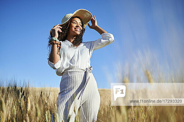 Lächelnde junge Frau mit Hut  die wegschaut  während sie in einem Weizenfeld steht
