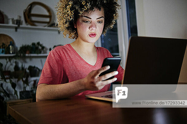Beautiful young woman sitting with laptop using smart phone at home