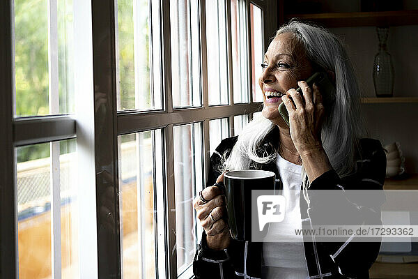 Happy woman with coffee mug looking through window while talking on smart phone at home