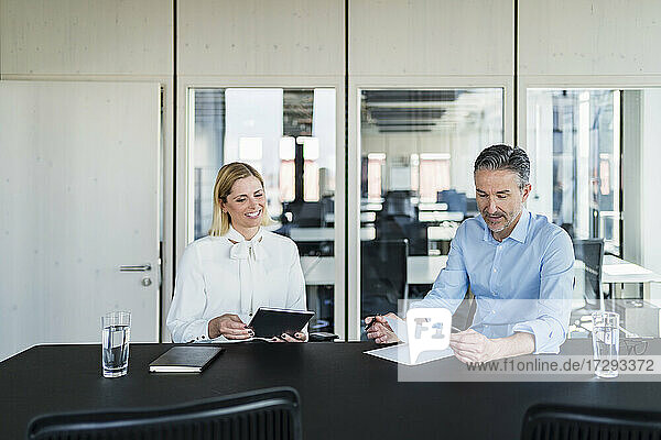Lächelnde Geschäftsfrau  die einen männlichen Kollegen beim Lesen von Dokumenten beobachtet  während sie am Schreibtisch im Büro sitzt