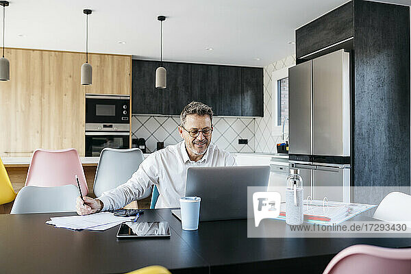 Lächelnder Geschäftsmann  der auf seinen Laptop schaut  während er in der Küche zu Hause arbeitet