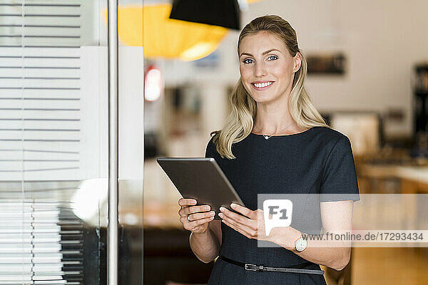 Lächelnde blonde Geschäftsfrau mit digitalem Tablet  die am Eingang eines Büros steht
