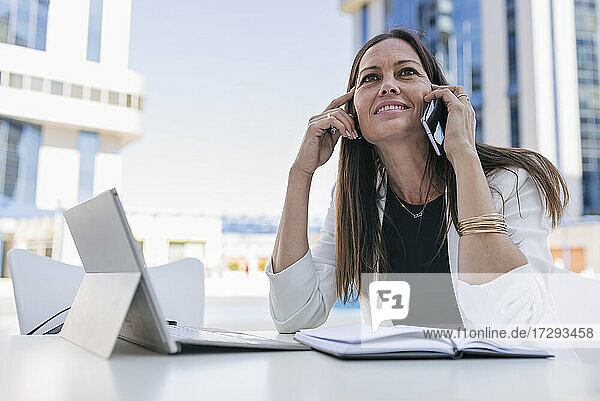 Lächelnde Geschäftsfrau mit digitalem Tablet  die in einem Straßencafé mit ihrem Mobiltelefon spricht