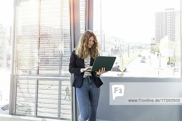 Weibliche Fachkraft  die einen Laptop benutzt  während sie vor einem Fenster im Büro steht