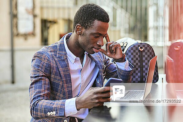 Junger männlicher Unternehmer mit Mobiltelefon und Laptop in einem Straßencafé