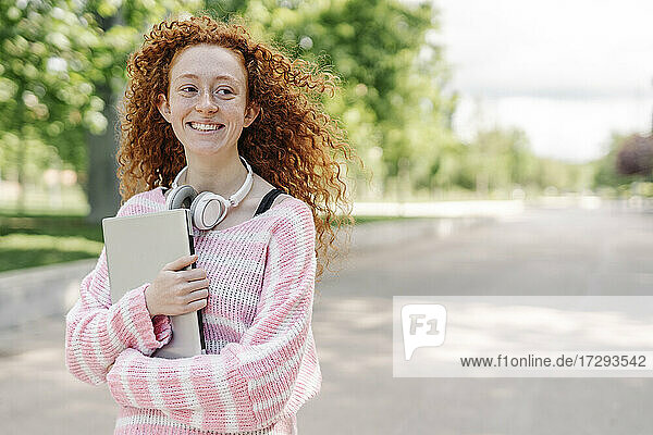 Glückliche junge Frau mit lockigem Haar trägt Laptop im Park