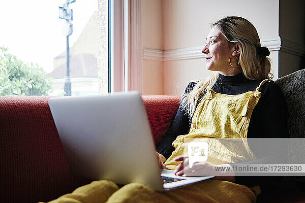 Nachdenkliche Frau mit Laptop  die wegschaut  während sie im Wohnzimmer sitzt