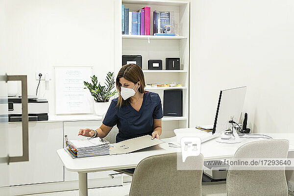 Zahnärztin mit Gesichtsschutzmaske  die am Schreibtisch in einer Klinik medizinische Unterlagen überprüft