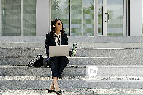 Geschäftsfrau mit Laptop  die wegschaut und einen Reisebecher hält  während sie auf einer Treppe sitzt