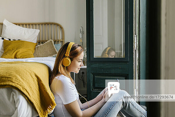 Lächelnde Frau mit Kopfhörern  die ein Smartphone benutzt  während sie sich zu Hause auf dem Bett anlehnt