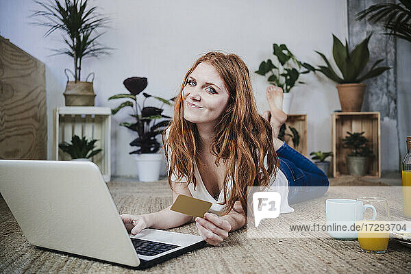 Lächelnde rothaarige Frau mit Kreditkarte beim Online-Shopping auf dem Laptop zu Hause