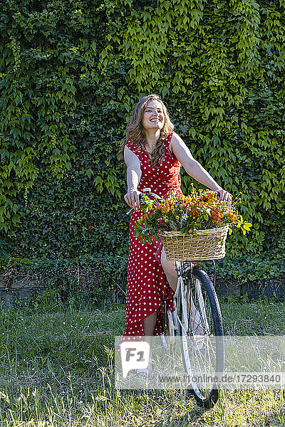 Glückliche junge Frau sitzt auf einem Fahrrad vor einer Hecke und schaut weg