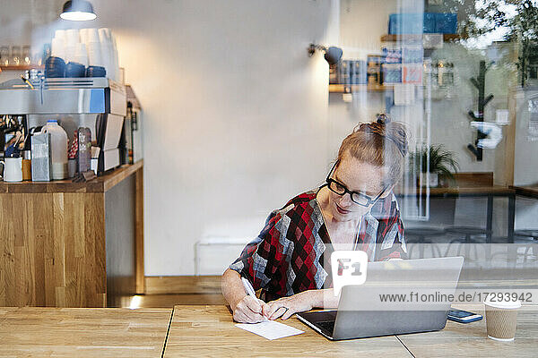 Frau schreibt auf Papier  während sie in einem Café vor einem Laptop sitzt
