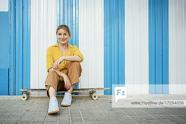 Beautiful woman sitting on longboard at footpath in front of striped wall