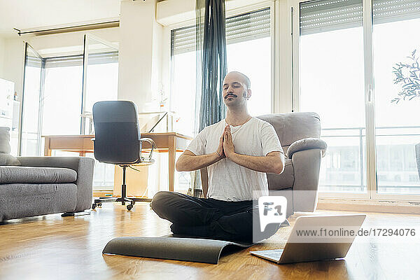 Mann mit verschränkten Händen meditiert auf einer Matte sitzend zu Hause
