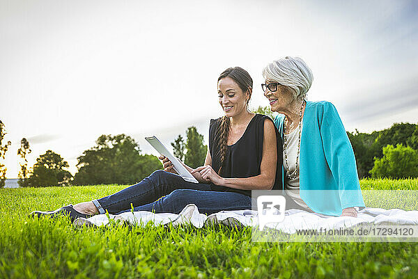 Smiling granddaughter using digital tablet while sitting by grandmother on picnic blanket