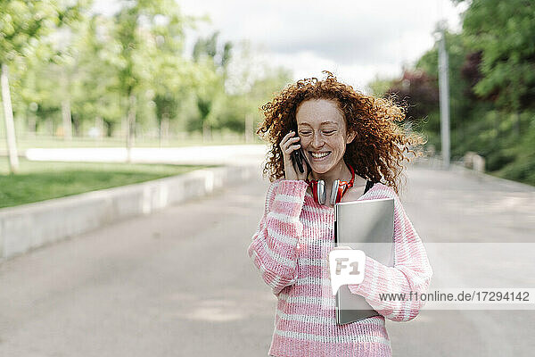Cheerful young woman carrying laptop while talking on mobile phone at park