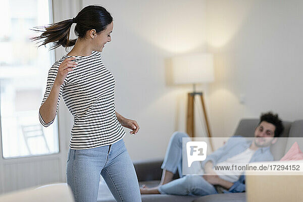 Junge Frau tanzt vor ihrem Freund  der sich zu Hause auf dem Sofa anlehnt