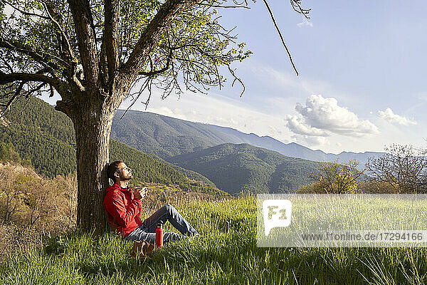 Mann mit roter Jacke lehnt an einem Baum und genießt die Natur