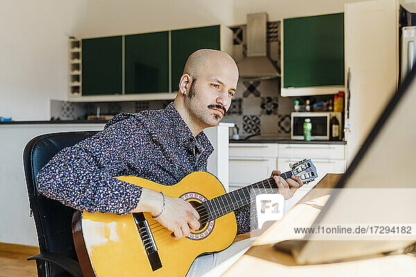 Männlicher Gitarrist schaut auf den Computer  während er im Wohnzimmer Gitarre spielt