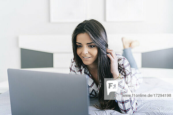 Frau mit Hand im Haar  die einen Laptop benutzt  während sie zu Hause auf dem Bett liegt