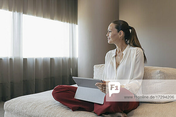 Female entrepreneur sitting cross-legged with digital tablet on sofa looking away