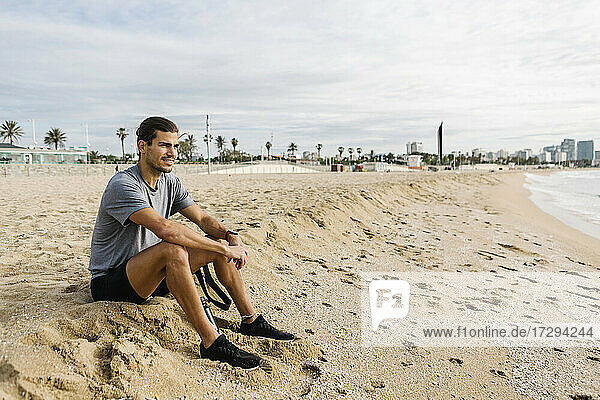 Männlicher Sportler schaut weg  während er am Strand im Sand sitzt