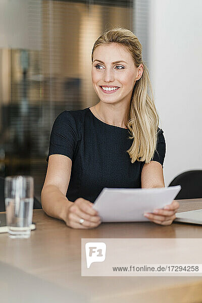 Beautiful smiling young entrepreneur sitting with document at desk while looking away