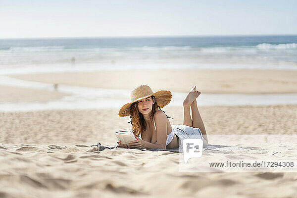 Junge Frau mit Buch  die wegschaut  während sie am Strand im Urlaub auf dem Sand liegt