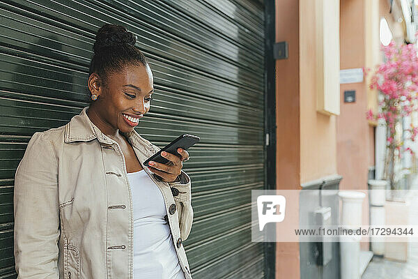 Schöne lächelnde Frau  die ein Smartphone benutzt  während sie vor einem Fensterladen steht